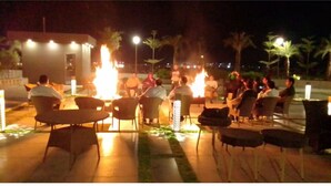Un espacio al aire libre con sillas dispuestas alrededor de una chimenea central, rodeado de palmeras y farolas.