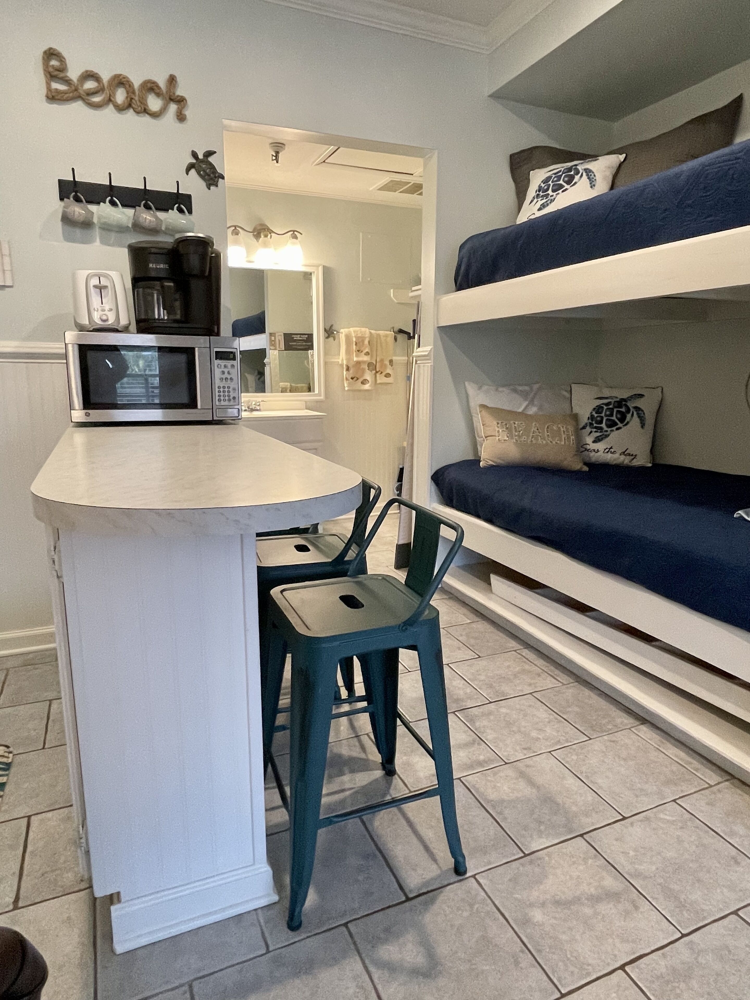 A compact kitchen with a microwave, a small island, and a chair. There is a bunk bed with pillows and a towel rack with a turtle decoration.