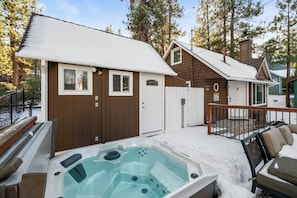 Un bain à remous avec une terrasse en bois, un bâtiment brun avec une porte blanche et une zone délimitée par une clôture.
