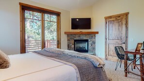 Une chambre avec un grand lit, une cheminée, un téléviseur fixé au-dessus de la cheminée et un balcon donnant sur la forêt.