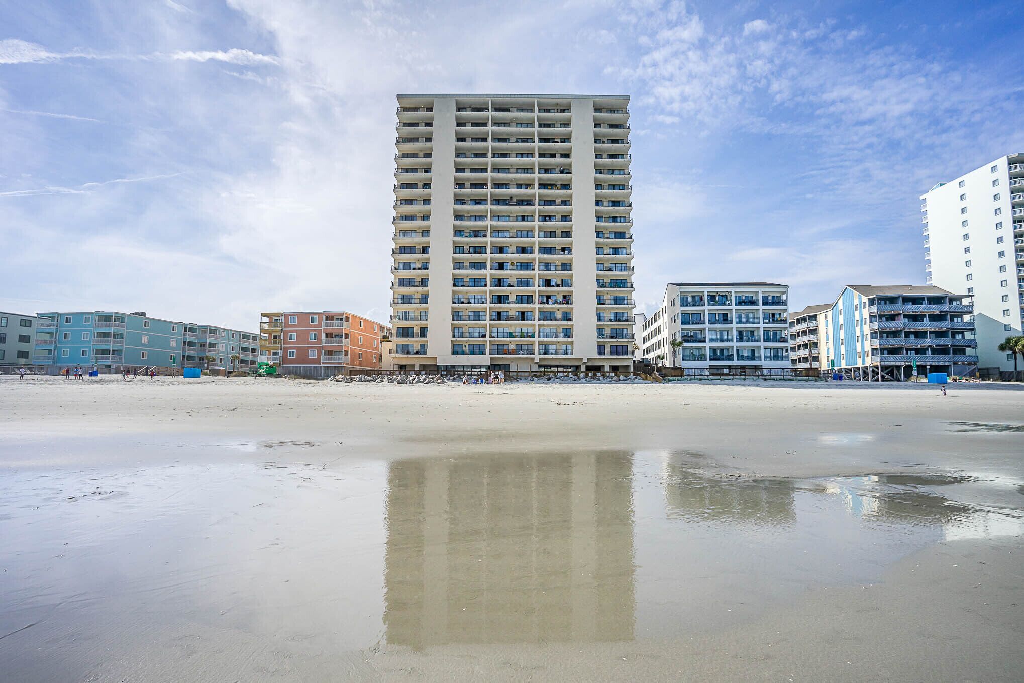 Ein Strandhotel mit einem spiegelnden Pool im Vordergrund, ein hohes Gebäude mit Balkonen und weitere Gebäude im Hintergrund.