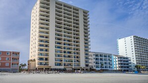 A tall, multi-story hotel with balconies, located near a beach.