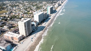 An aerial view of a coastal city with a long beachfront, high-rise buildings, and a clear sky.
