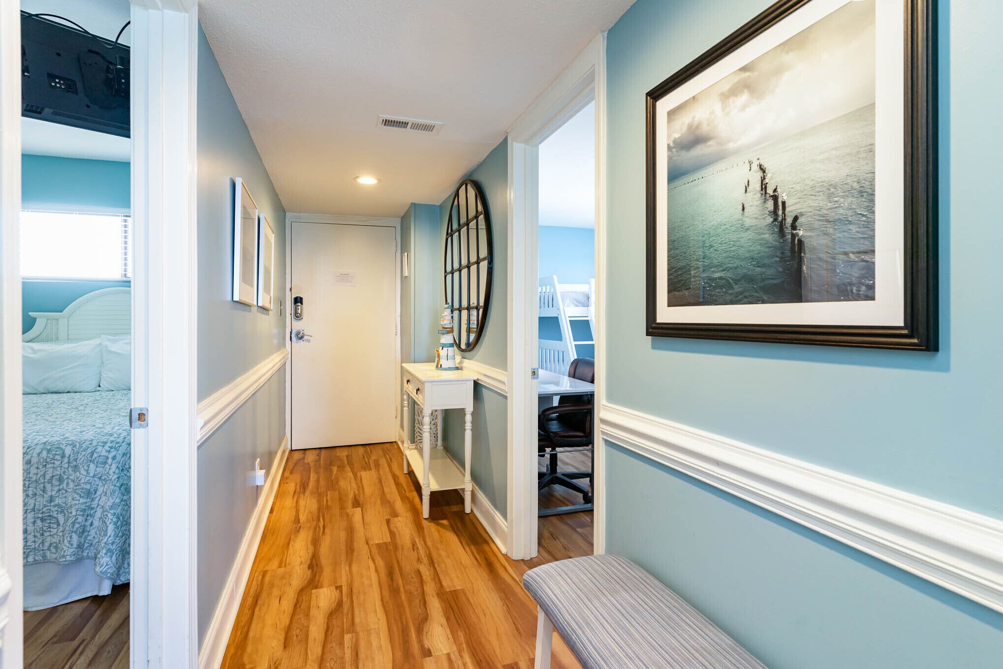 A hallway with wooden flooring, a white bench, a desk with a chair, and a framed picture on the wall.