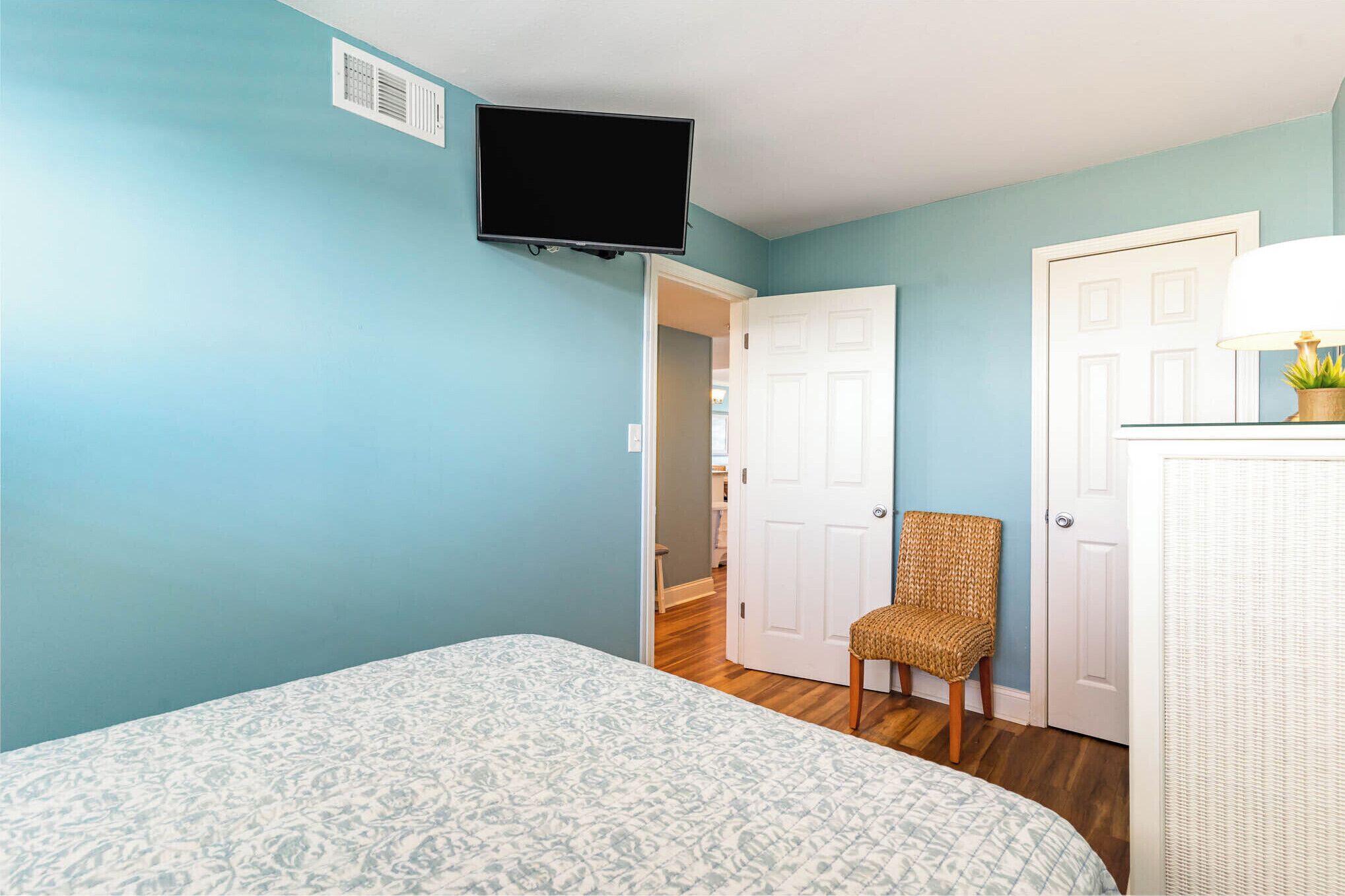Une chambre à coucher comprenant un lit, un téléviseur fixé au mur, un fauteuil en osier et une porte donnant sur une autre pièce.