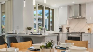 Una cocina moderna con armarios blancos, una campana extractora de acero inoxidable y una zona de comedor con una mesa preparada para cuatro.