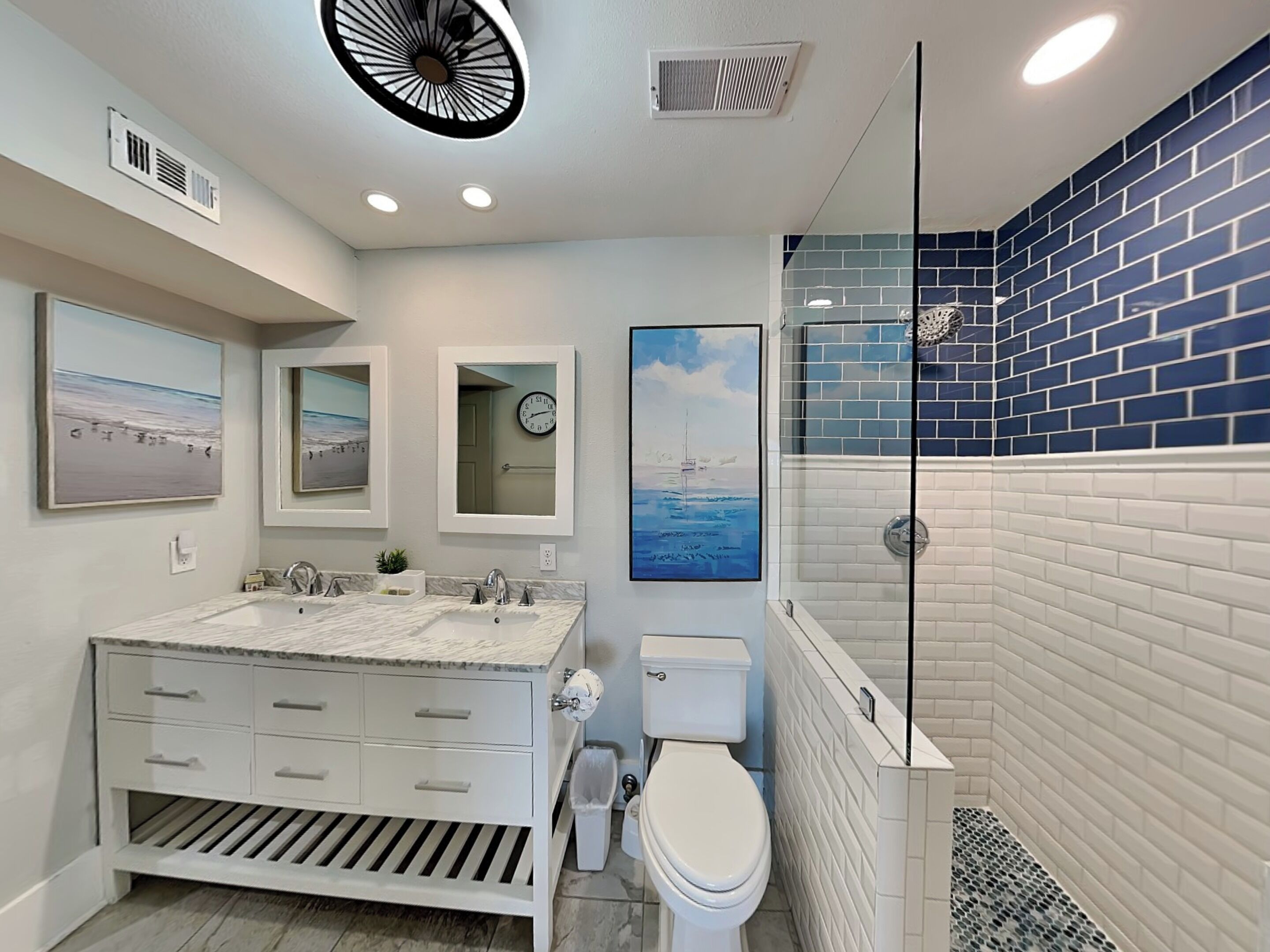 A bathroom with a white vanity, a toilet, a shower with blue tiles, and artwork on the walls.