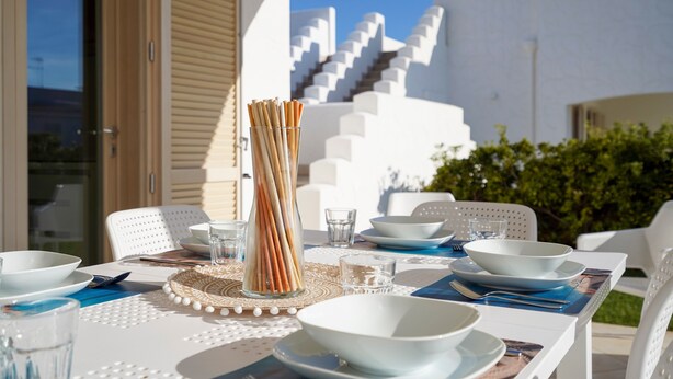 Un espace extérieur aménagé pour les repas, avec des meubles blancs, un vase en verre contenant des branches, et une vue sur des bâtiments blancs et des escaliers.