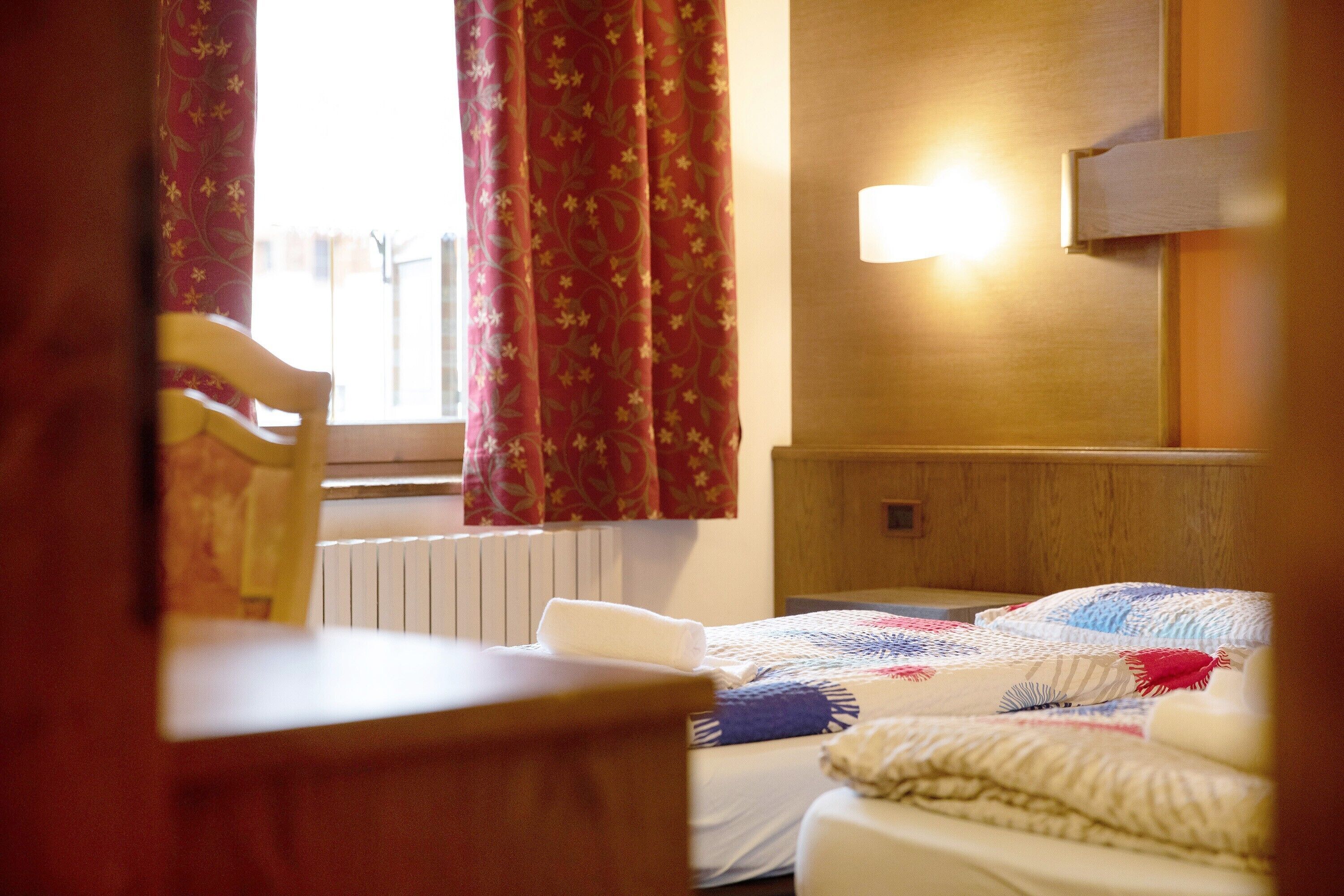 A hotel room with a bed, a chair, a window with curtains, and a wall-mounted light.
