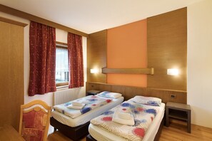 A hotel room with two beds, a wooden headboard, a window with curtains, a wall-mounted light, and a small table.
