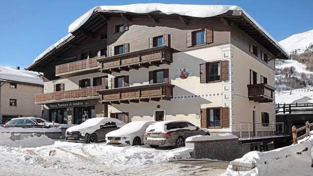 A multi-story building with a snow-covered roof, a balcony with wooden railings, and a sign indicating a bank. - Apartment "Casa San Marco N 1" with Mountain View, Garden & Wi-Fi (Livigno)