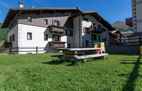 A traditional chalet with a stone bench, wooden deck, and a mountain backdrop.