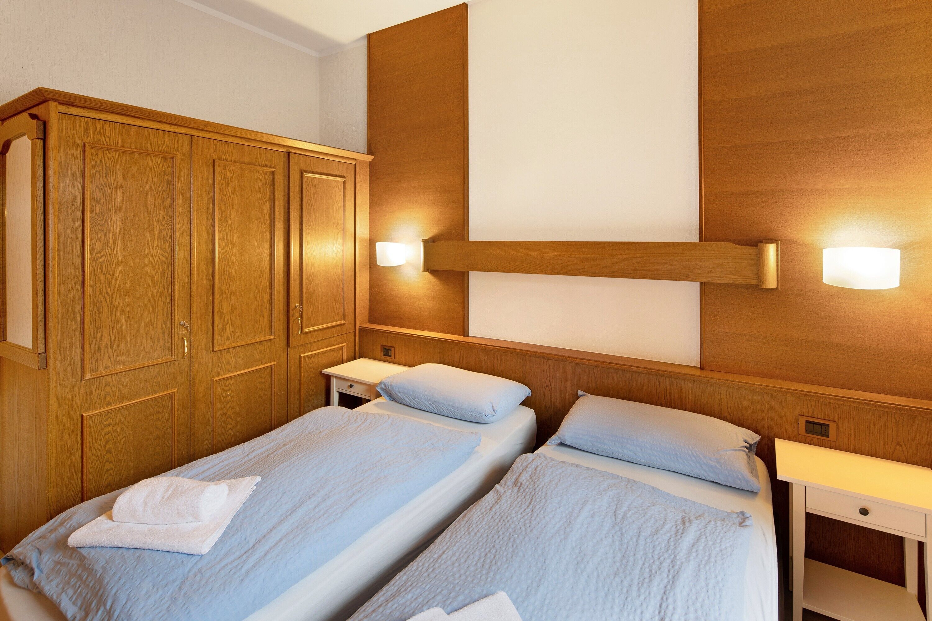 Une chambre d’hôtel avec deux lits, une armoire en bois, des tables de chevet et des lampes fixées au mur.