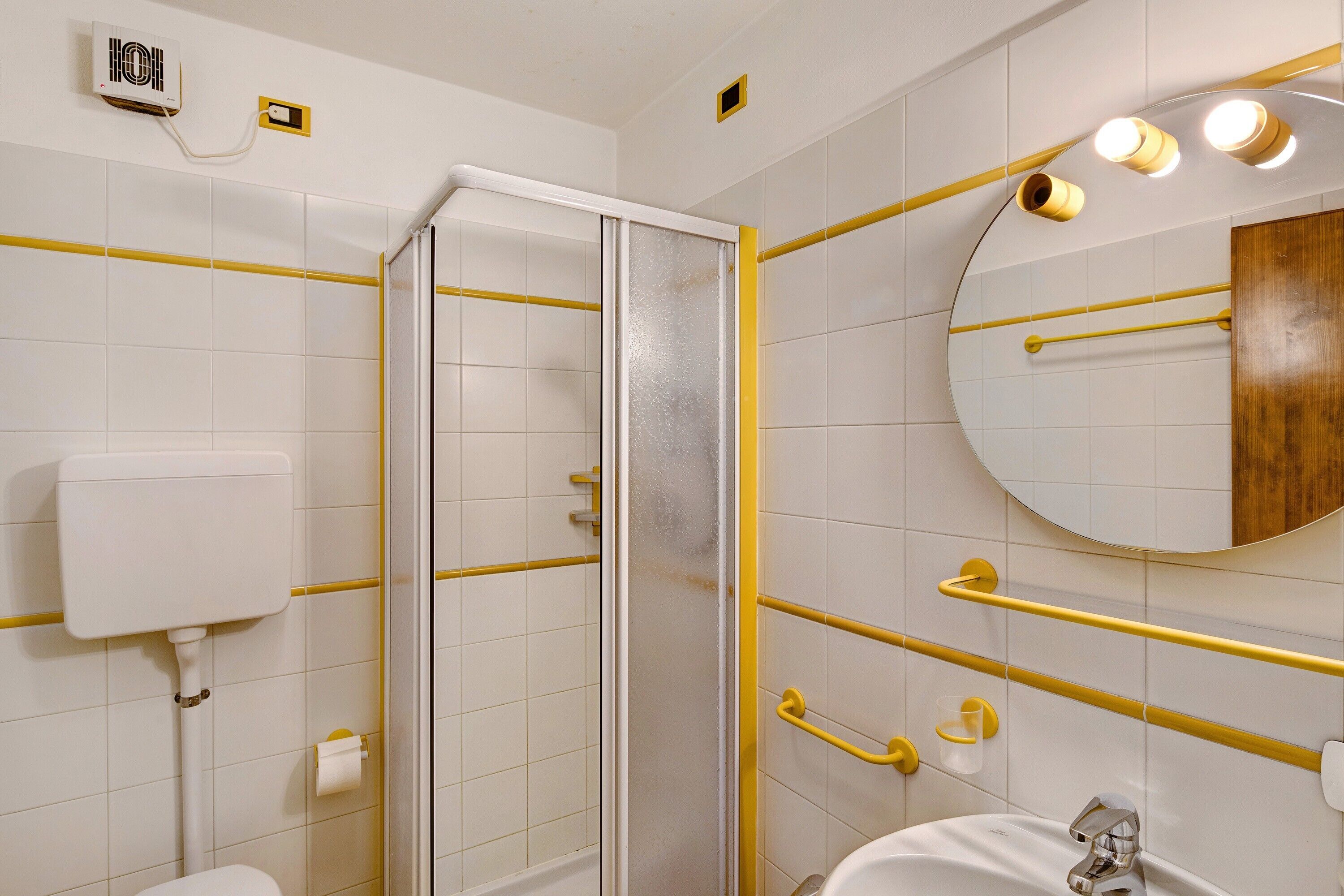 A bathroom with a toilet, a sink, a mirror, a shower with glass doors, and yellow safety bars.