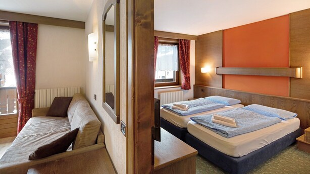 Ein Zimmer mit einem Bett, einer Schlafcouch, einem Fenster mit Vorhängen und einer Wandleuchte.