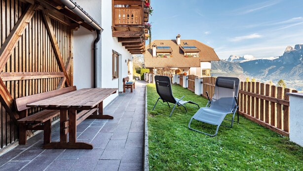 Een houten gevel met een bank en stoelen, een terras en uitzicht op bergen.