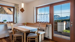 Een eethoek met een houten tafel en stoelen, een raam met uitzicht op bergen, en een radiator onder het raam.