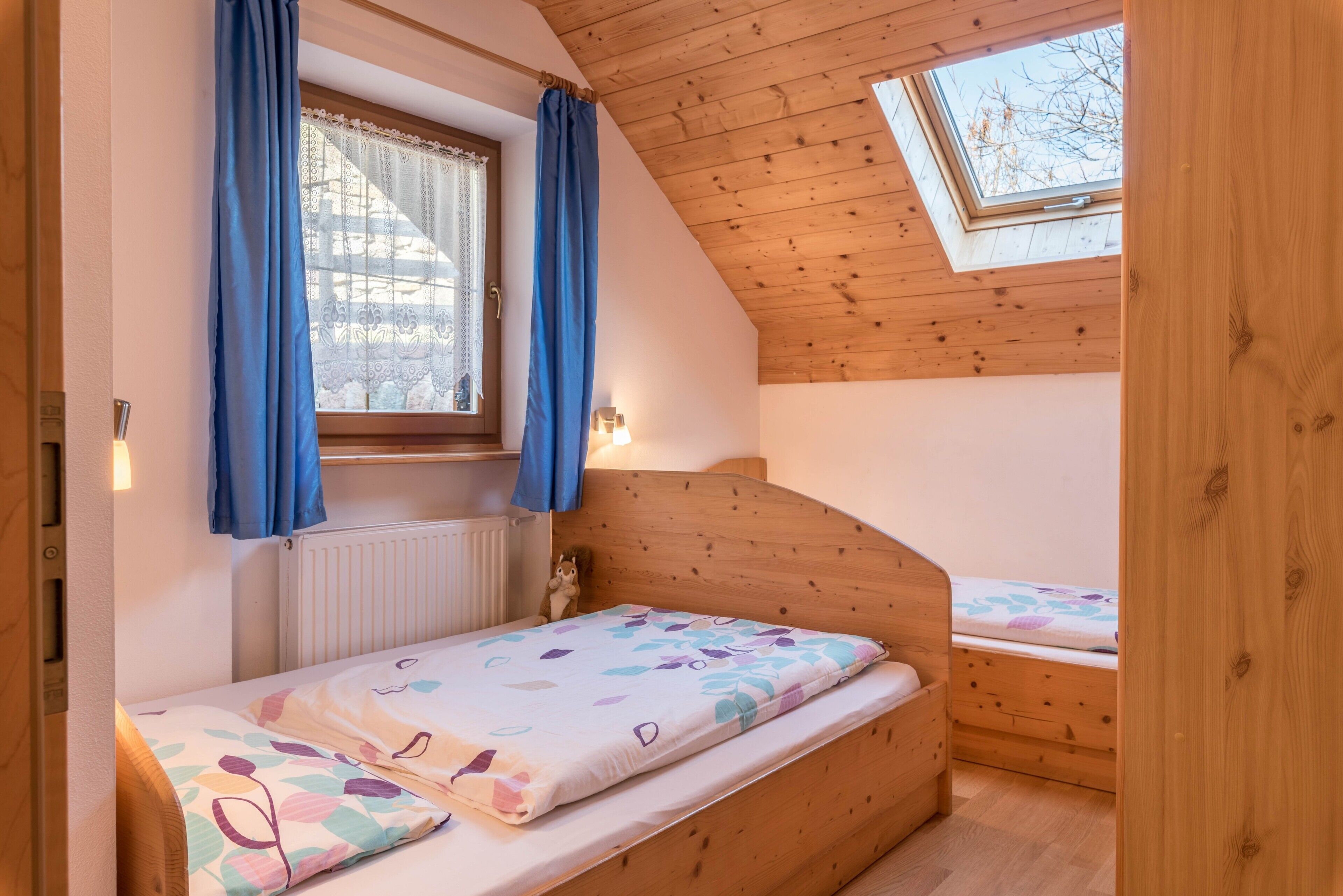 Une petite pièce agréable, avec un plafond en pente, une fenêtre avec des rideaux, un lit recouvert d’une courtepointe à motifs floraux et une petite commode.