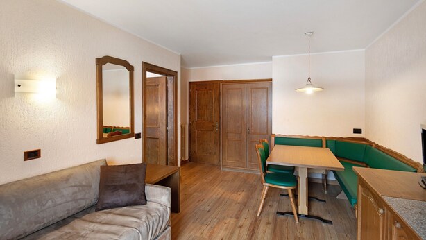 Ein Zimmer mit Holzboden, einer Couch, einem Esstisch mit Stühlen, einer grünen Bank und Holztüren.