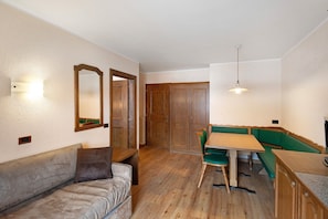 A room with a wooden floor, a sofa, a dining table with chairs, a green bench, and wooden doors.