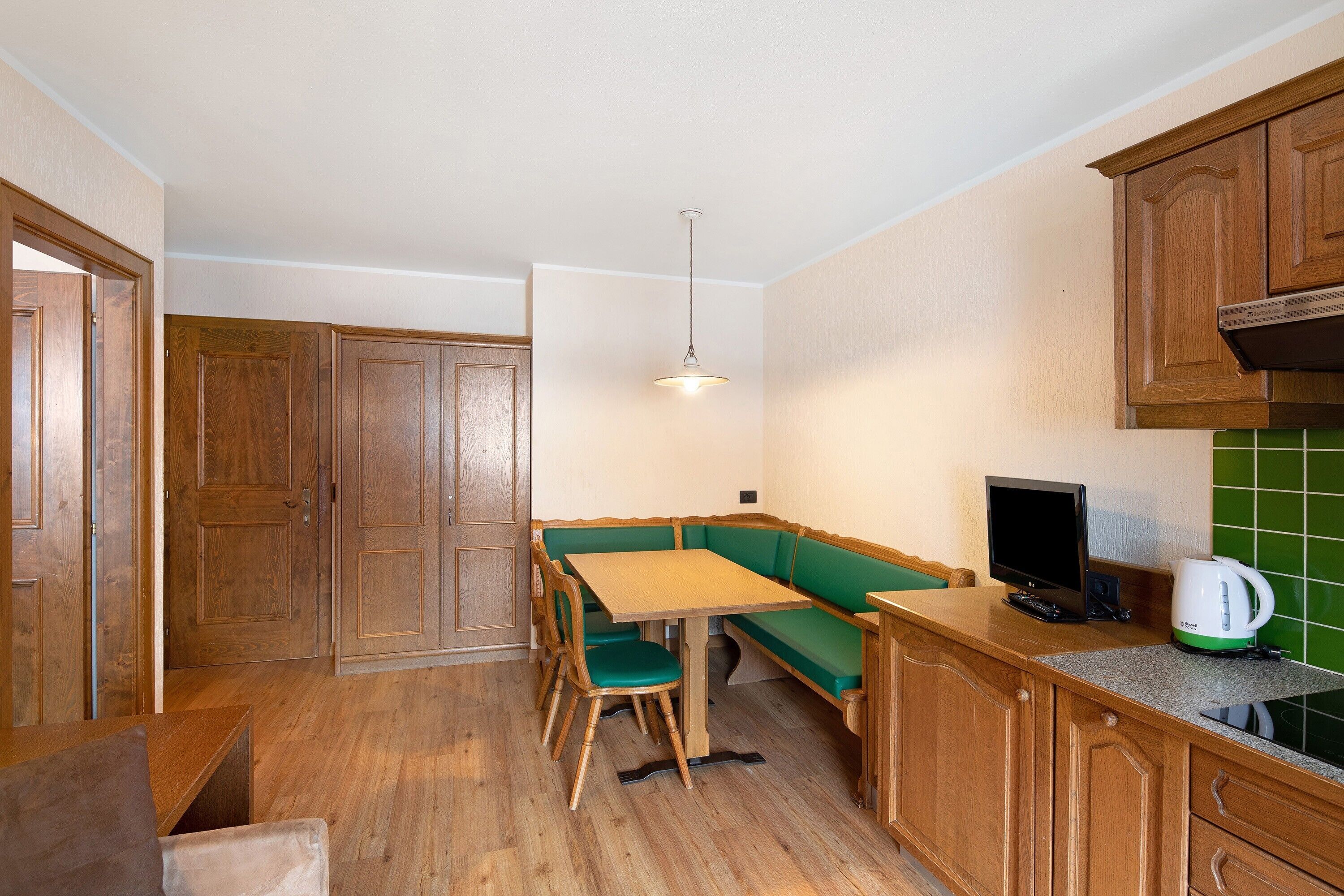 A compact kitchen with wooden cabinets, a green bench, a wooden table, and a kettle on the counter.