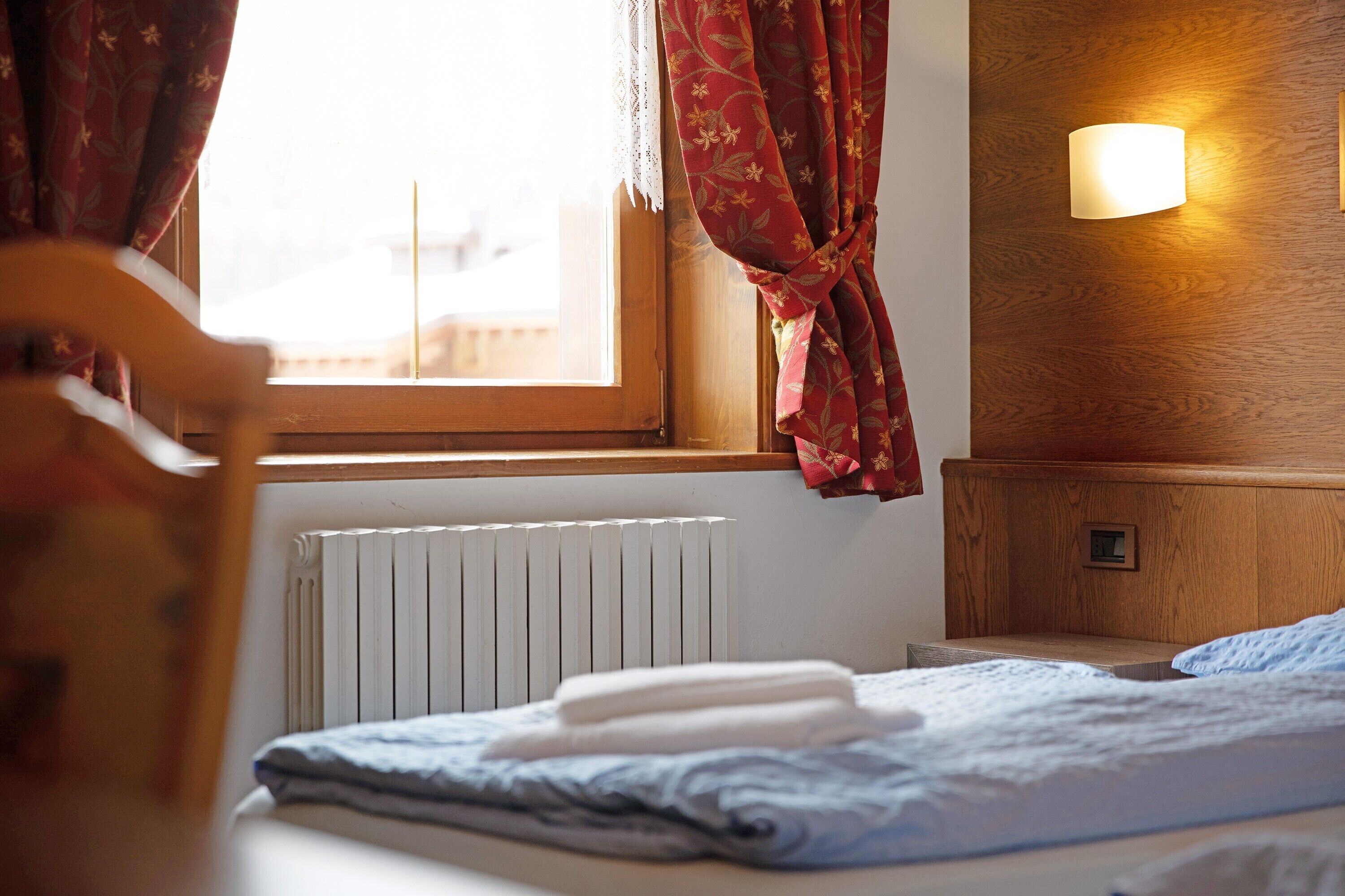 A bedroom with a bed, a window with curtains, a wall-mounted light, and a radiator.