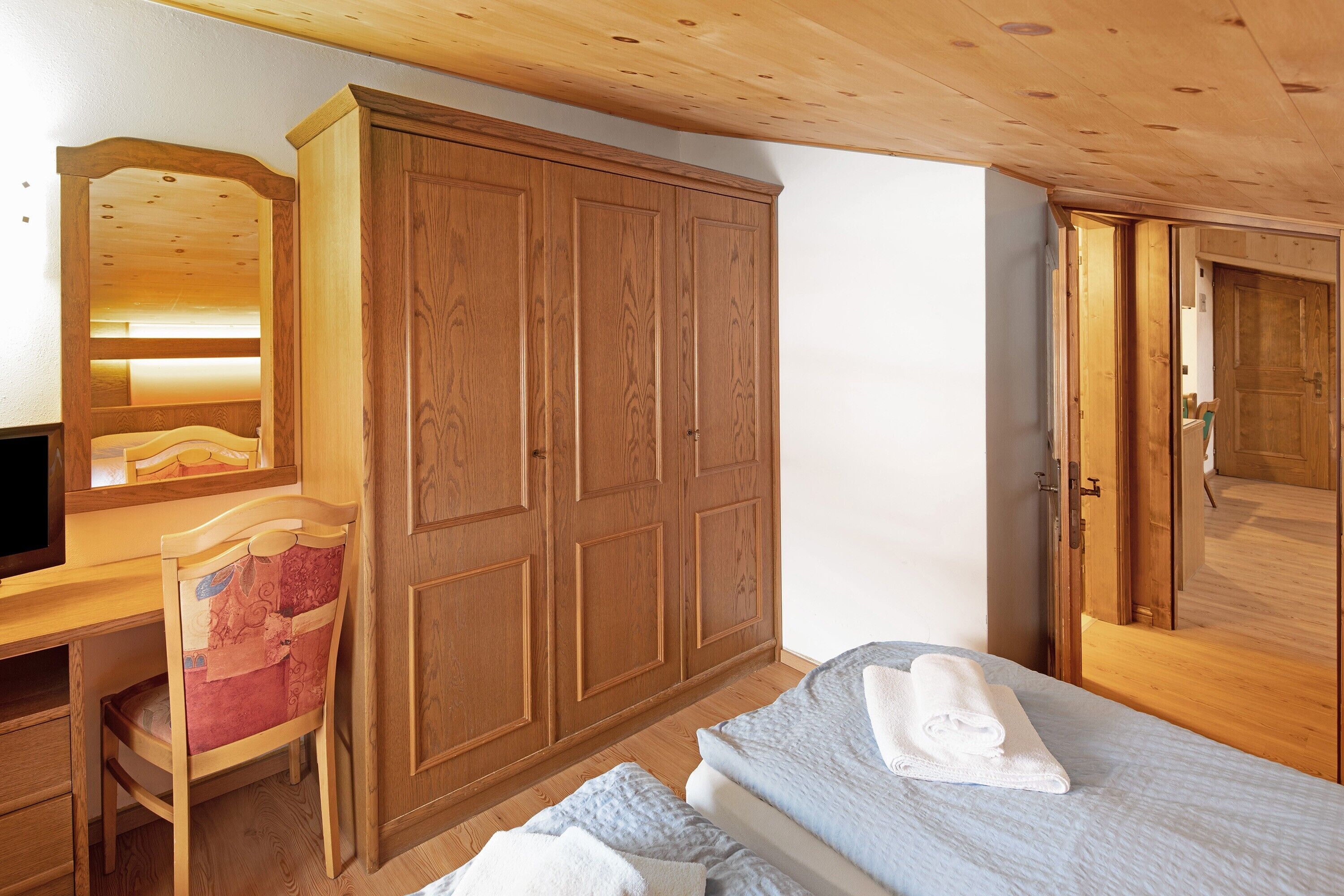 Une chambre avec une armoire en bois, un bureau avec une chaise et un lit sur lequel est posée une serviette enroulée.