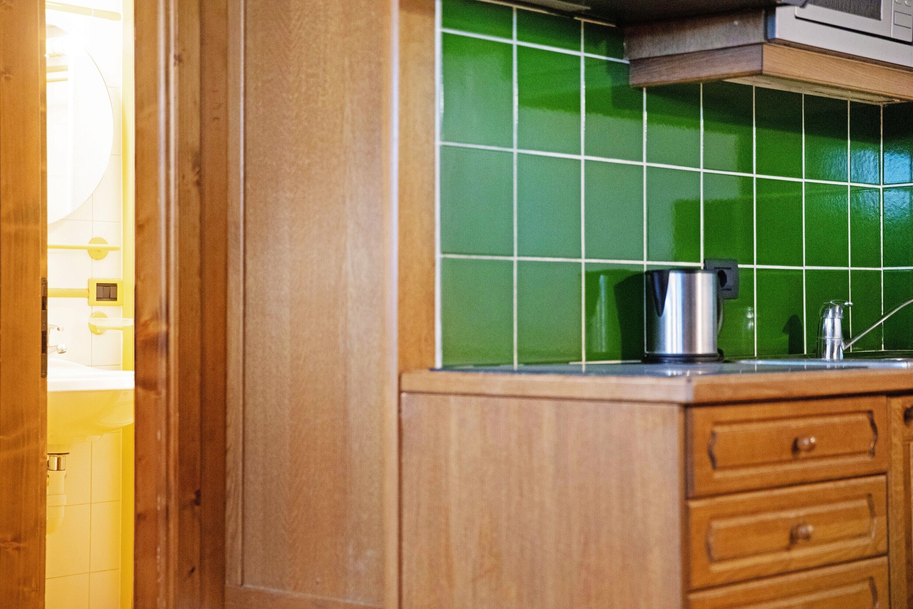 A kitchen with green tiles, wooden cabinets, and a stainless steel kettle.