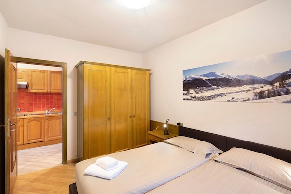 Une chambre à coucher avec un lit, une table de chevet, une armoire en bois et une cuisine visible à travers une porte ouverte.