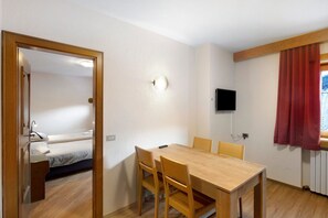 A room with a wooden dining table, four chairs, a TV mounted on the wall, and a bed in the adjacent room.