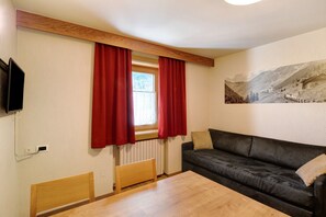 A room with a wooden headboard bed, a black leather sofa, a wooden dining table, a window with red curtains, and a wall-mounted TV.
