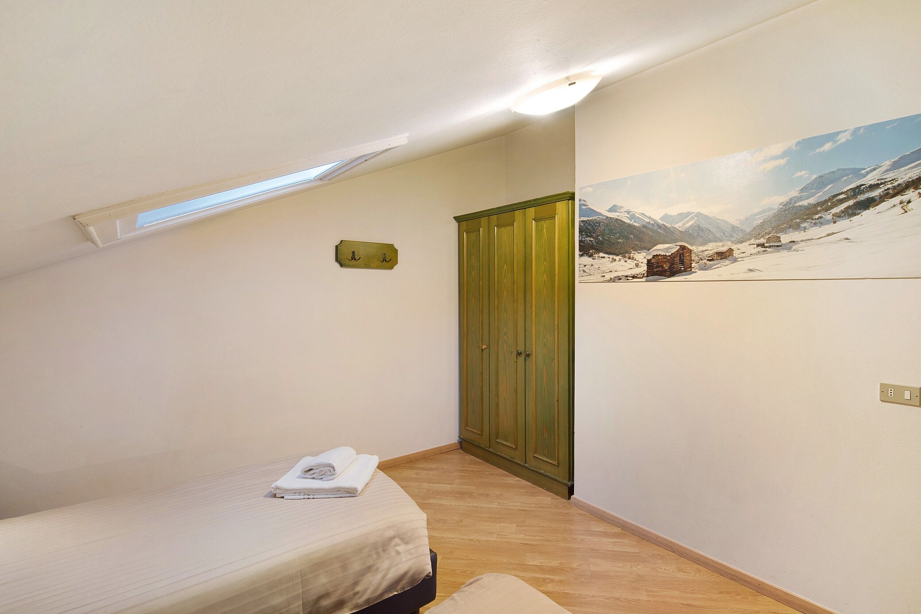 Une chambre à coucher avec un lit, une armoire, un tableau représentant un paysage enneigé et une lucarne.