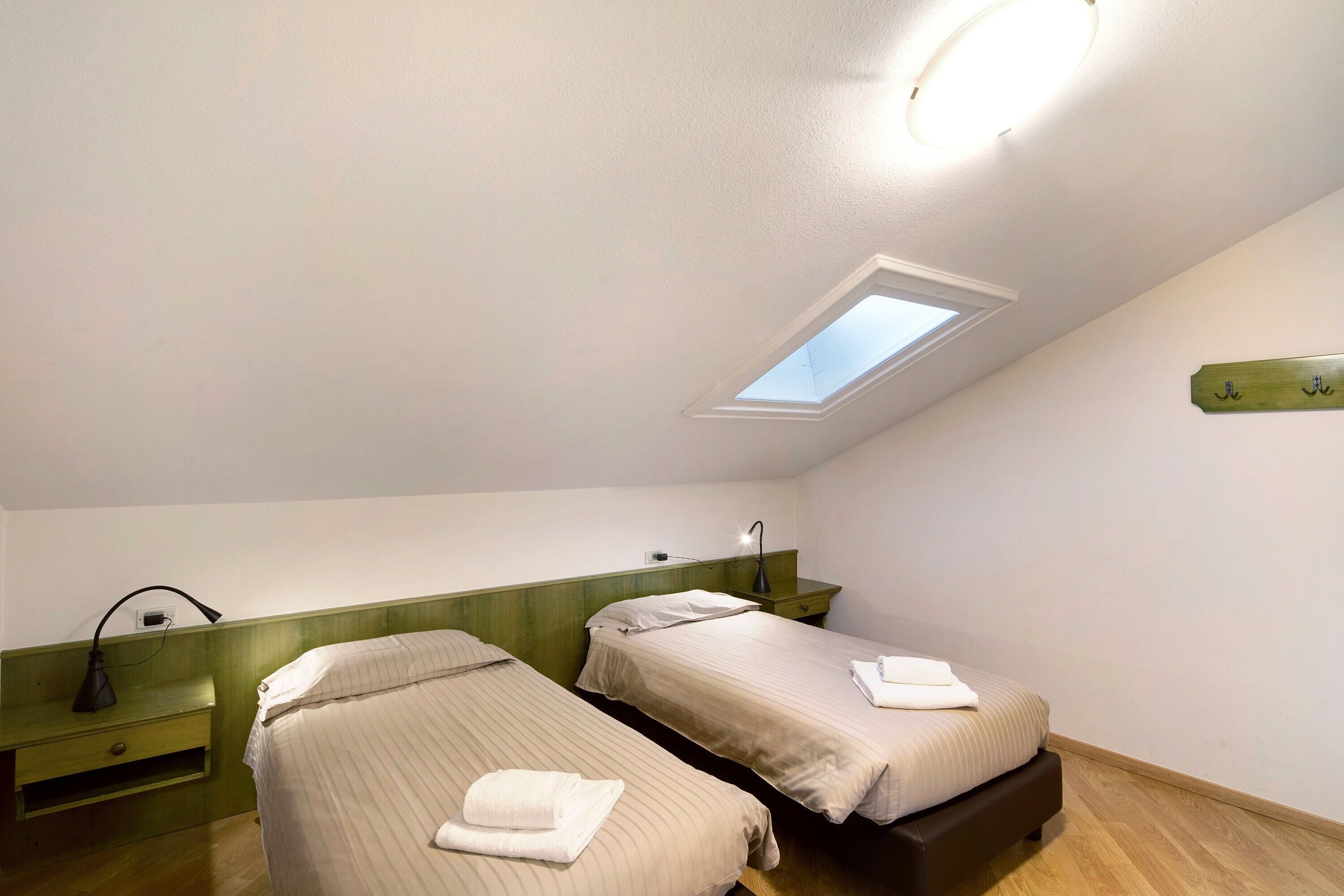 A room with two beds, a skylight, a bedside table with a lamp, and a wall-mounted shelf.