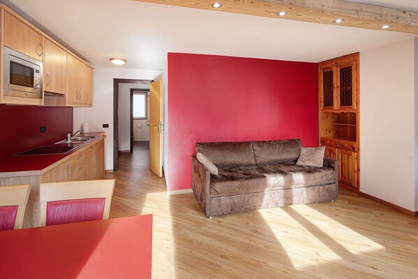 A compact living space with a kitchenette, a red accent wall, a brown leather sofa, and wooden flooring.