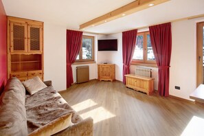 A room with a wooden floor, a sofa, a wooden cabinet, a TV mounted on the wall, and windows with curtains.