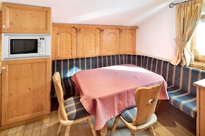 A corner room with a wooden cabinet, a microwave, a table with a pink tablecloth, striped bench seating, and a window with curtains.