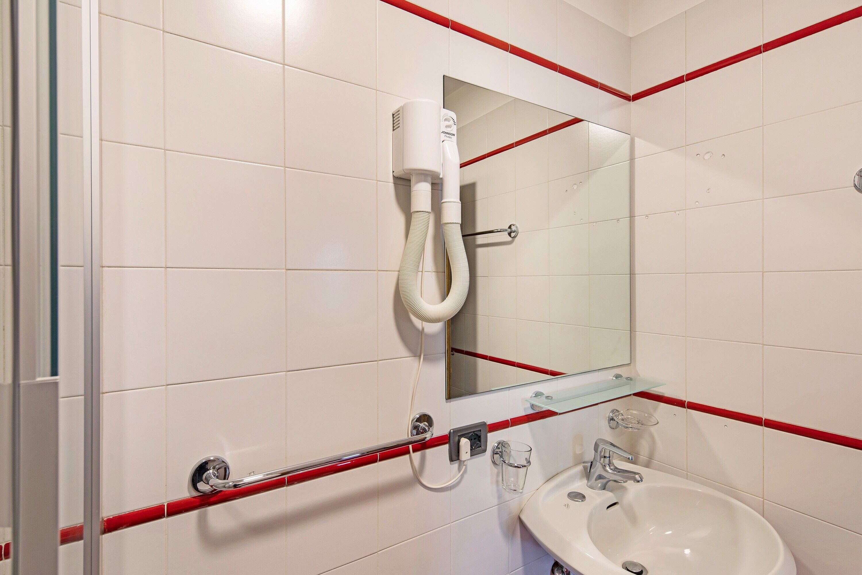 Une salle de bain avec des murs recouverts de carreaux blancs, une douche en verre, un lavabo blanc et des touches de rouge sur les murs et les éléments de décoration.