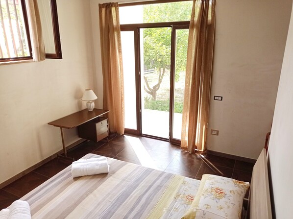 A bedroom with a bed, a desk, a lamp, and a view of greenery outside.