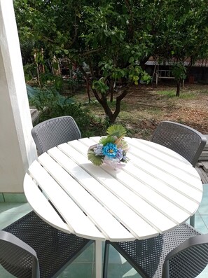A round white table with a floral centerpiece, surrounded by black mesh chairs, set on a balcony with a view of a garden and trees.