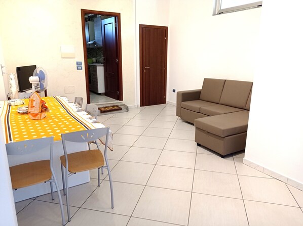 Un comedor con una mesa para dos personas, un sofá y una cocina visible a través de una puerta abierta.