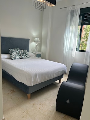 A bedroom with a bed, a nightstand, a lamp, a chair, and a window with curtains.