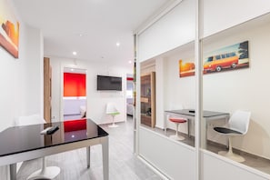 A modern hotel room with a glass partition, a flat-screen TV, a red accent wall, and a dining area with a table and chairs.