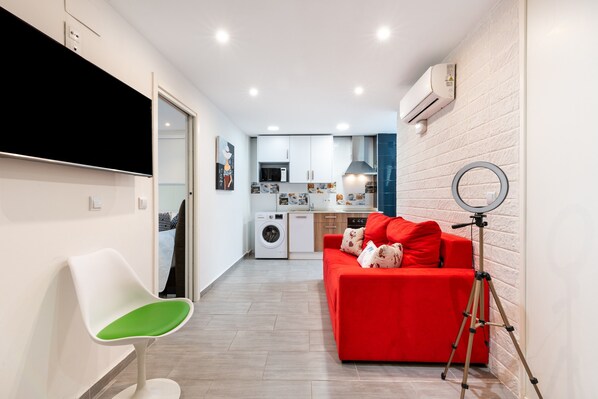 A modern living room with a red sofa, a white chair, a tripod, and a kitchen in the background.