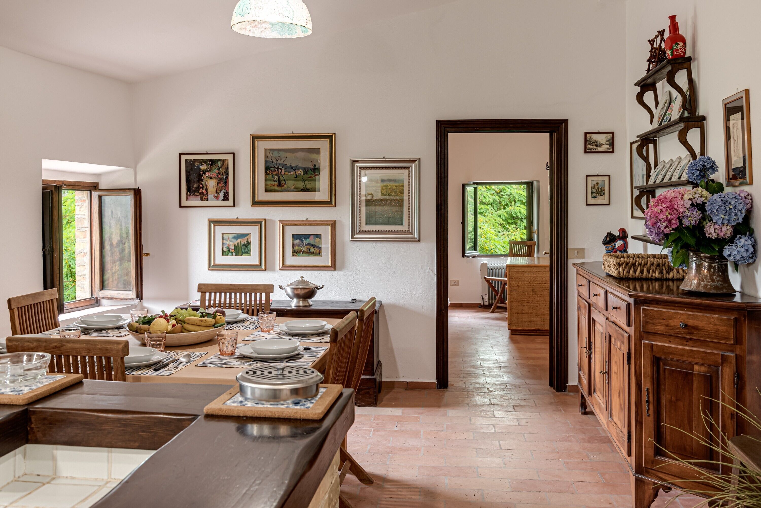 A dining area with a long wooden table set for a meal, surrounded by chairs. There are framed pictures on the walls, a wooden cabinet with decorative items, and a view of greenery outside.