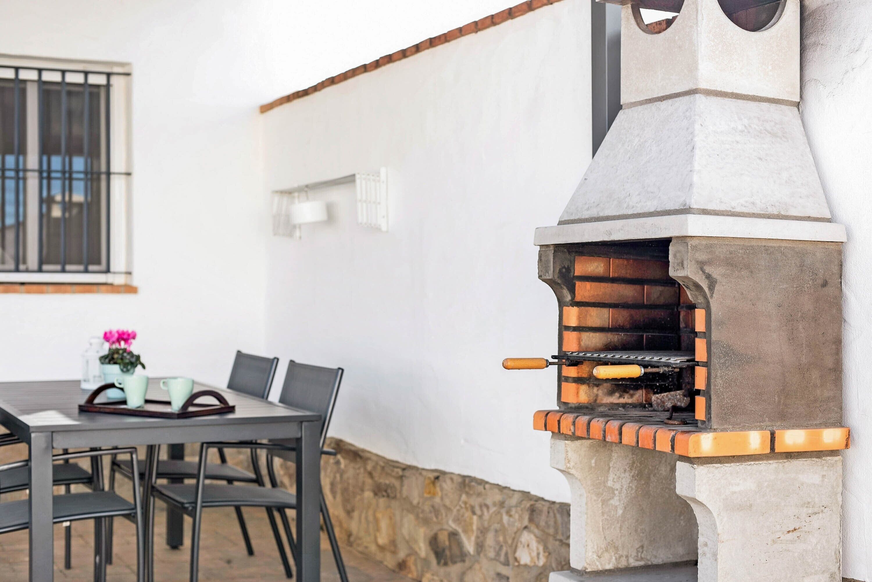 A stone-built outdoor oven with a chimney, a table with chairs, and a window with bars.