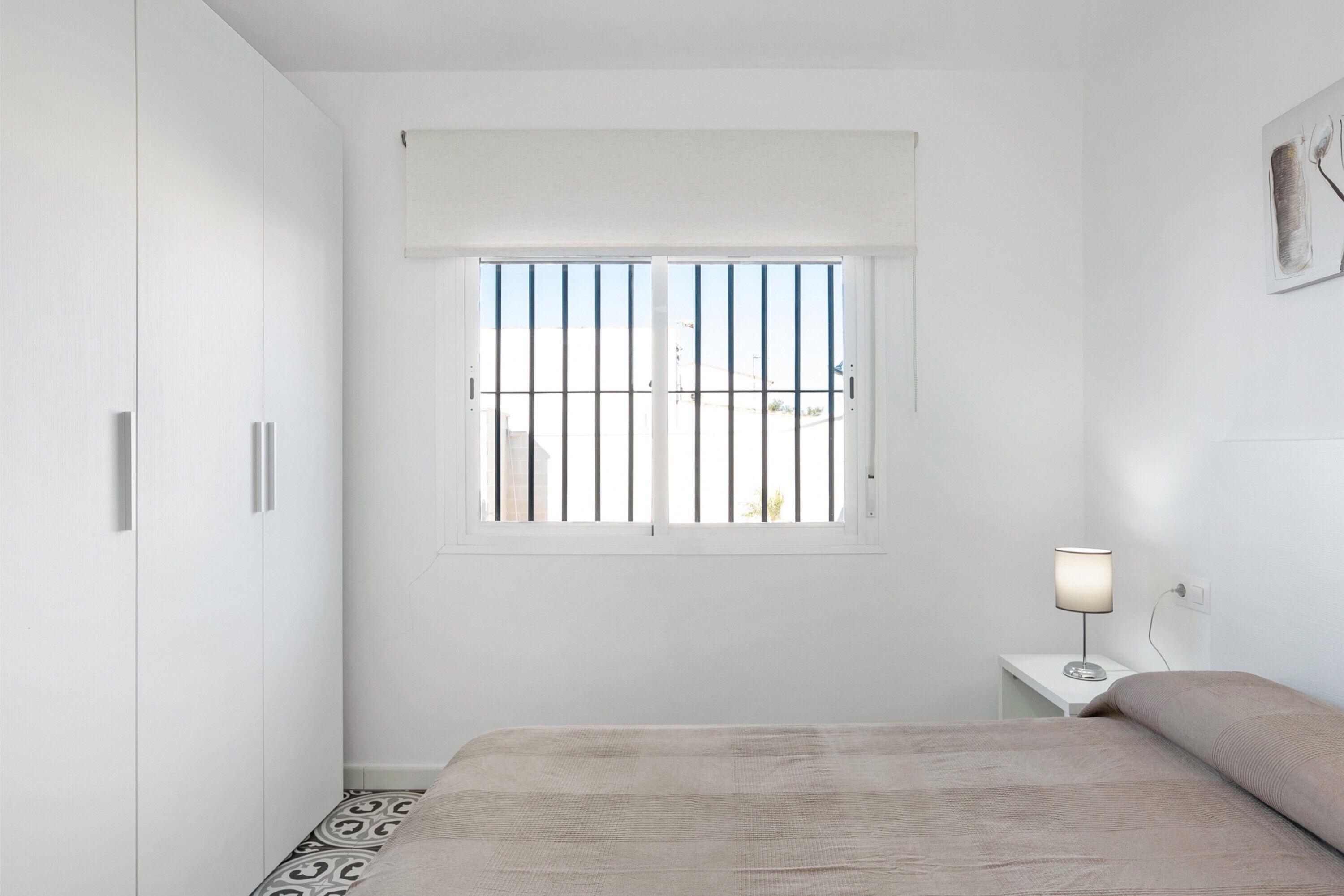 A bedroom with a bed, a nightstand, a lamp, a window with bars, and a wardrobe.