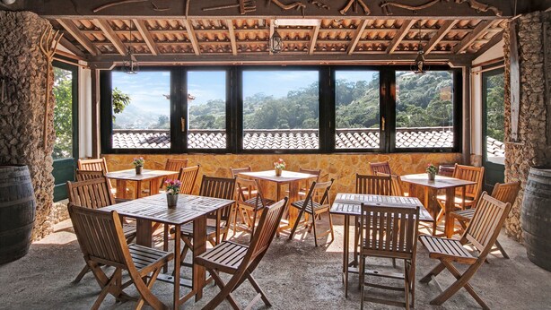 An outdoor dining area with wooden tables and chairs, a stone wall, and a view of a mountainous landscape through large windows. - Holiday Home "La Escalincha" with Mountain View, Shared Pool & Wi-Fi (Tegueste)