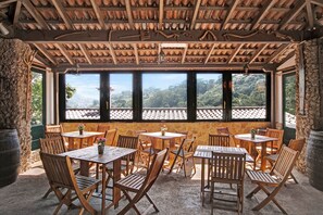 An outdoor dining area with wooden tables and chairs, a stone wall, and a view of a mountainous landscape through large windows.