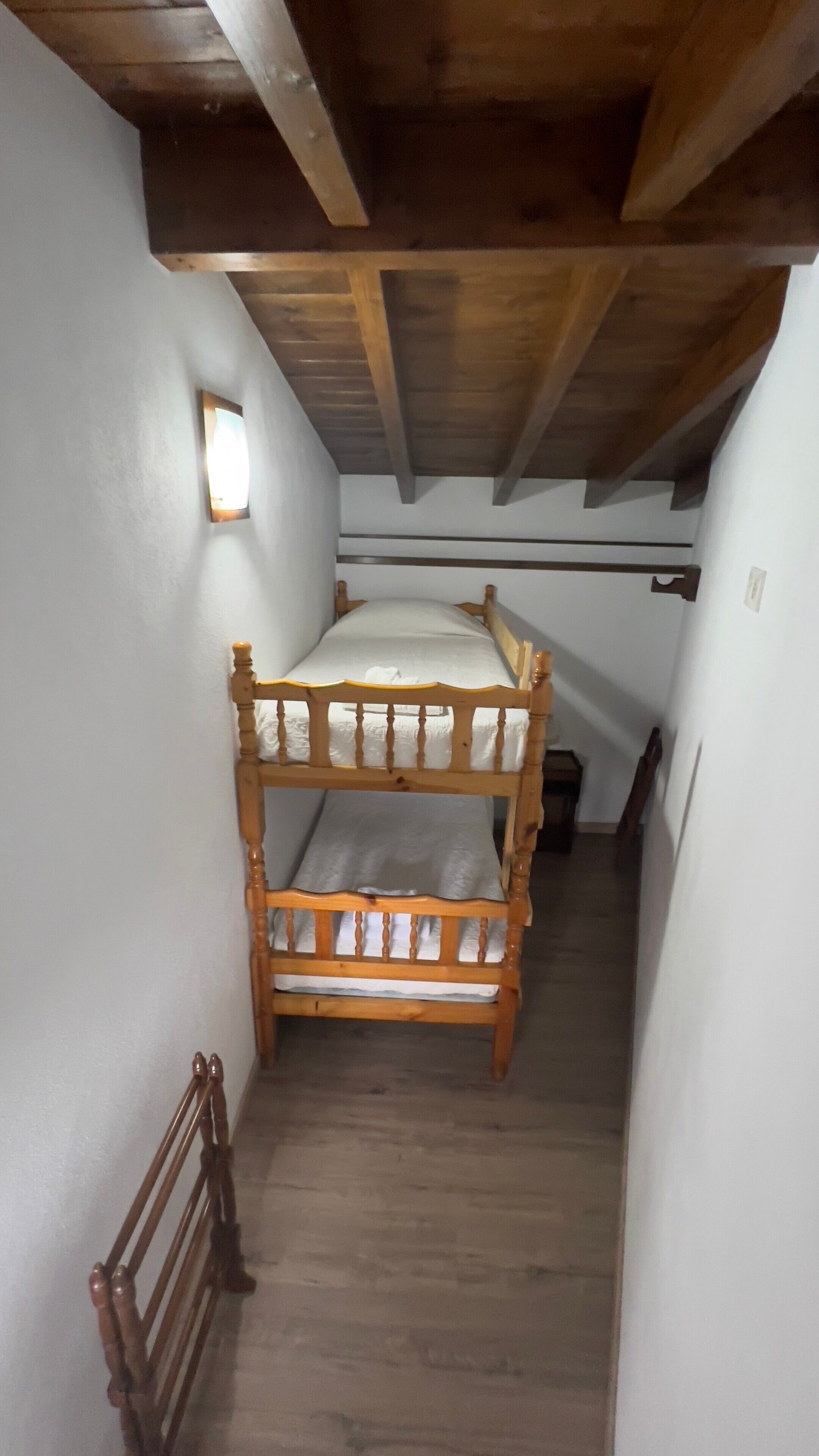 Una habitación estrecha con una cama litera de madera, un ventanal pequeño y una viga de techo de madera.