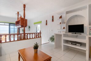 Un salon avec une table en bois, un téléviseur posé sur un meuble blanc, un balcon doté d’une balustrade et une vue sur la mer.